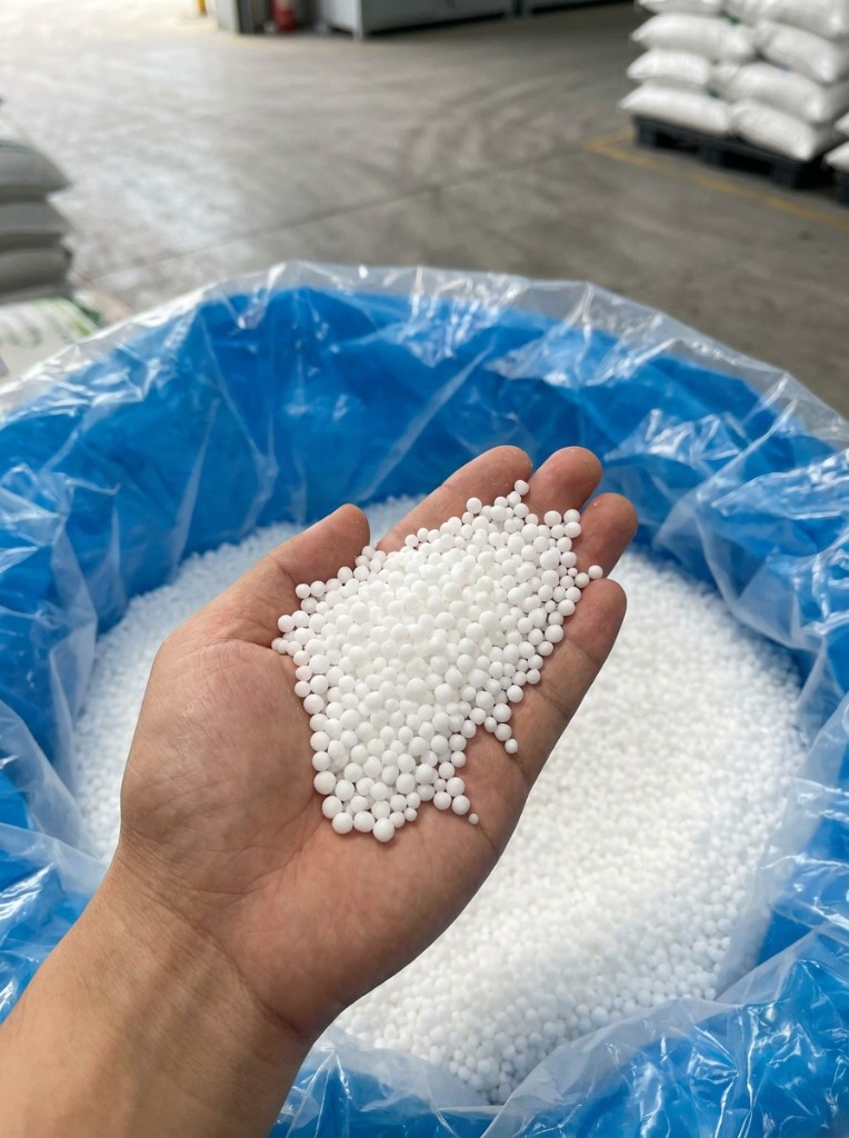 Urea prills quality check — sample in hand above bulk-lined tote with warehouse FIBC storage